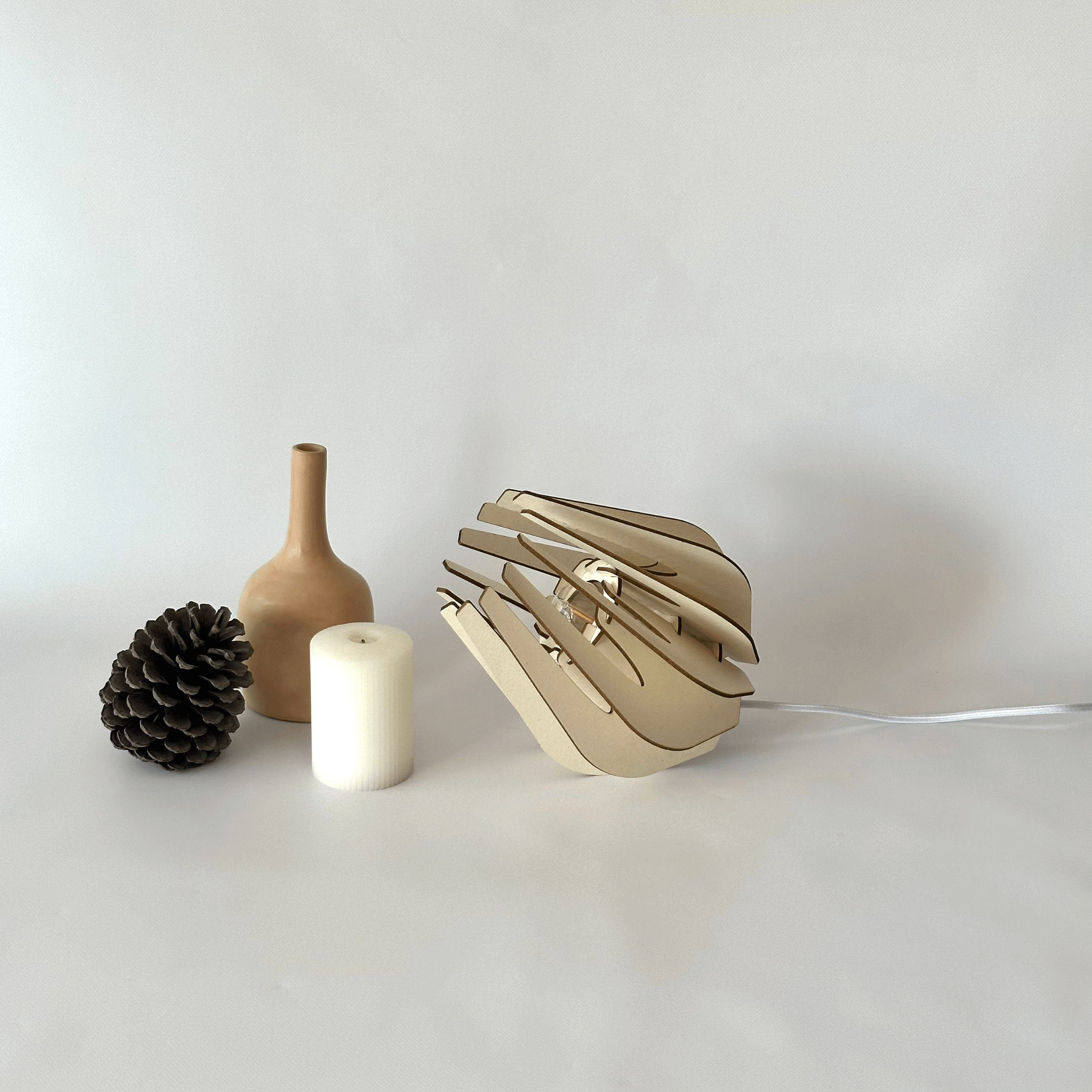 Le luminaire à Lucien mis en valeur durant un shooting photo sur fond blanc avec lumière naturelle. A droite un vase beige, une pigne de pin et une bougie accompagnent le luminaire.