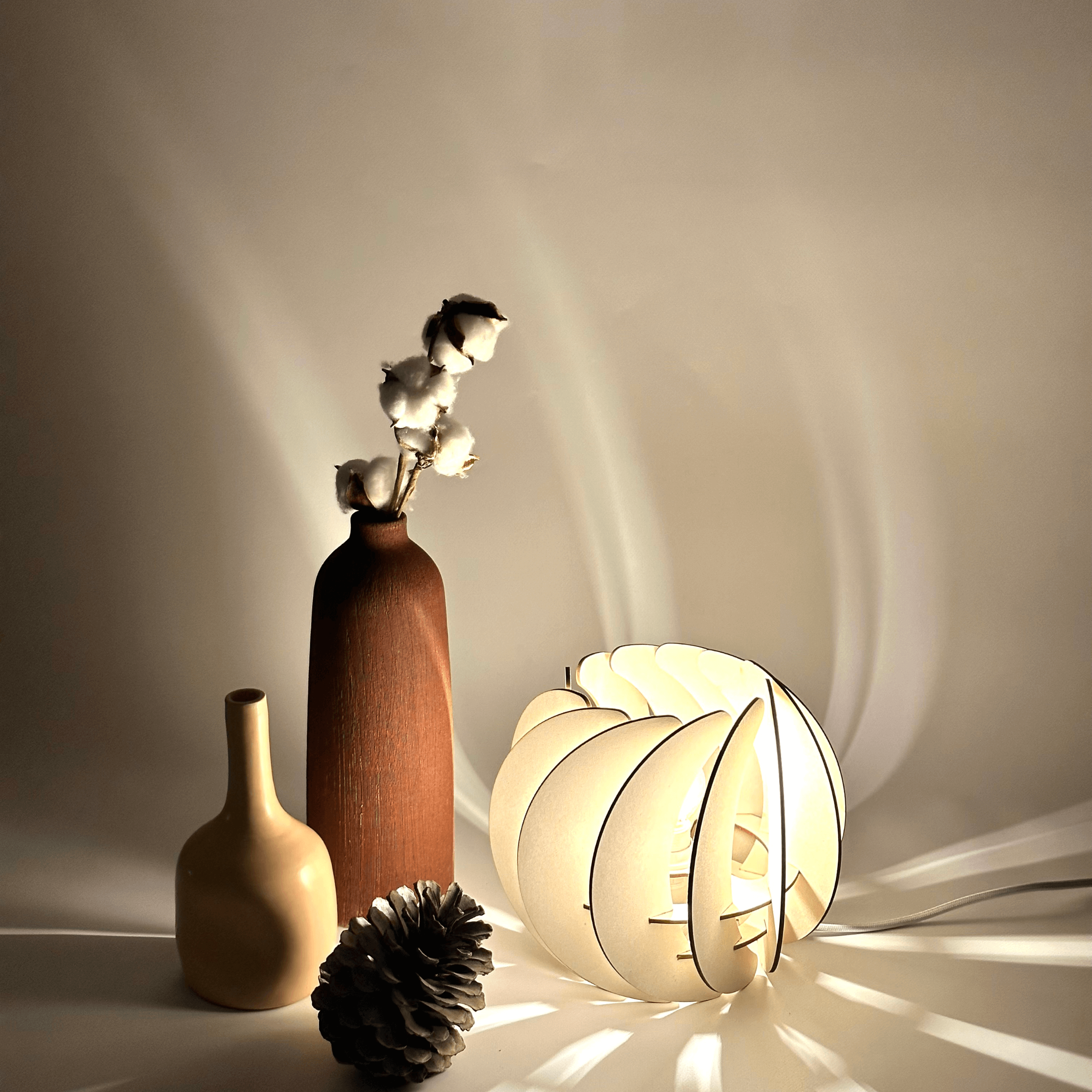 Luminaire Garance mis en valeur durant un shooting photo dans le noire. Garance dégage de somptueux éclat de lumière de manière symétrique. A droite de vase de couleur marron et brun avec un brin de coton et pigne de pin. 