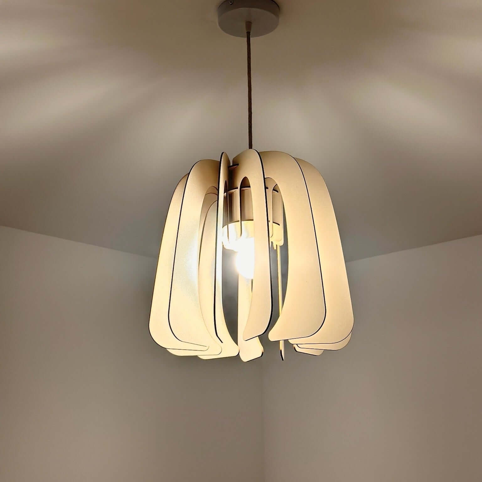 Un luminaire suspendu allumé, diffusant une lumière chaleureuse et douce. Son design sculptural est constitué de lames incurvées en matériau beige clair, qui créent un jeu d'ombres et de reflets subtils sur le plafond. Le câble en textile et la fixation discrète au plafond accentuent son style minimaliste et moderne, idéal pour éclairer avec élégance un intérieur épuré.