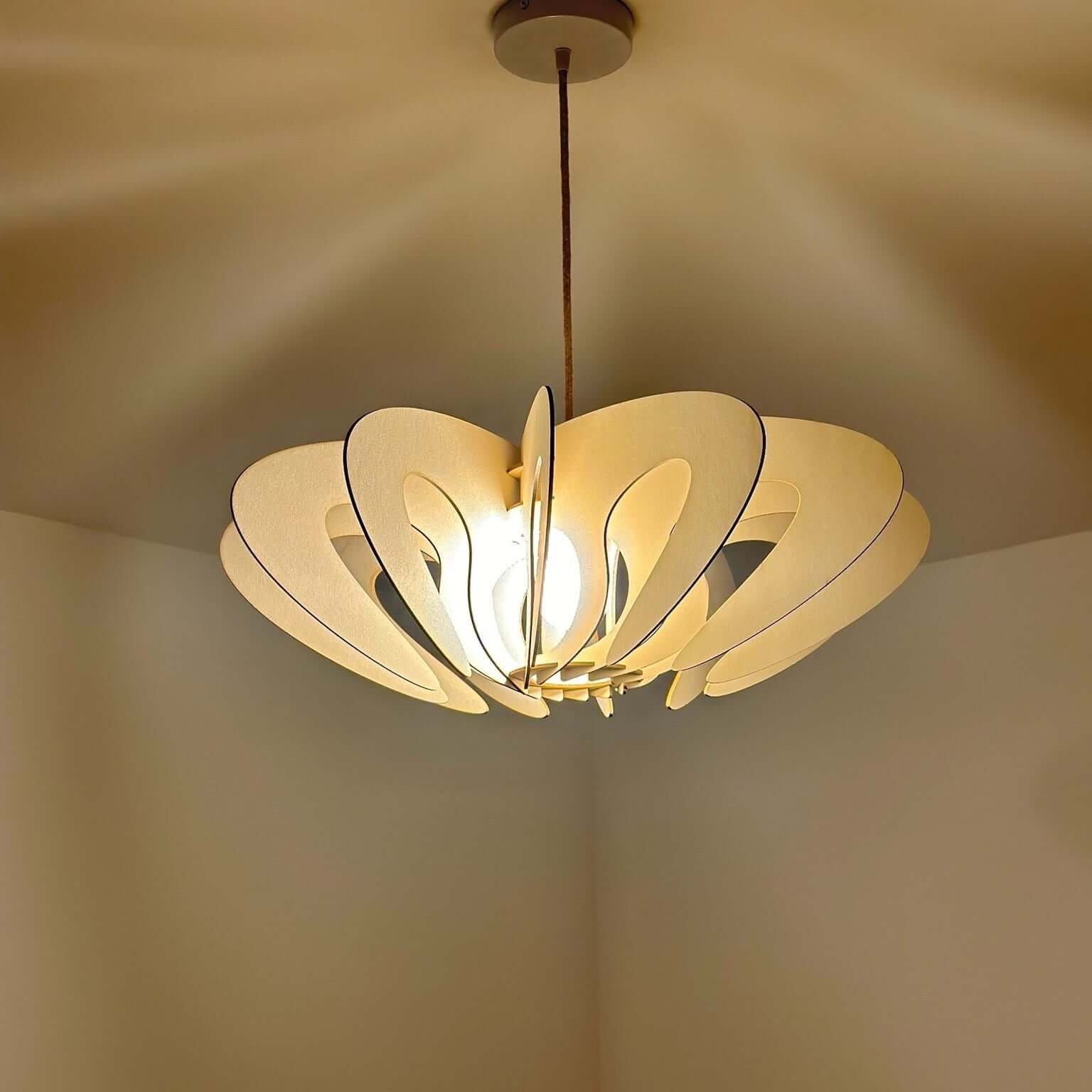 Ce luminaire suspendu en bois présente un design organique, avec des panneaux courbés et ajourés qui diffusent la lumière de manière élégante. Lorsqu’il est allumé, l’ampoule au centre projette des motifs subtils sur le plafond et les murs, créant une ambiance chaleureuse et accueillante. Les tons doux et naturels du bois sont mis en valeur par l’éclairage, ce qui accentue le caractère artisanal et moderne de cet objet décoratif.